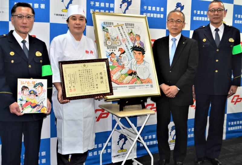 完成した防犯ポスターの前で、署長感謝状を受け取ったさとうさん（左から２人目）。多忙な連載の合間を縫い、すし職人の衣装で駆け付けた