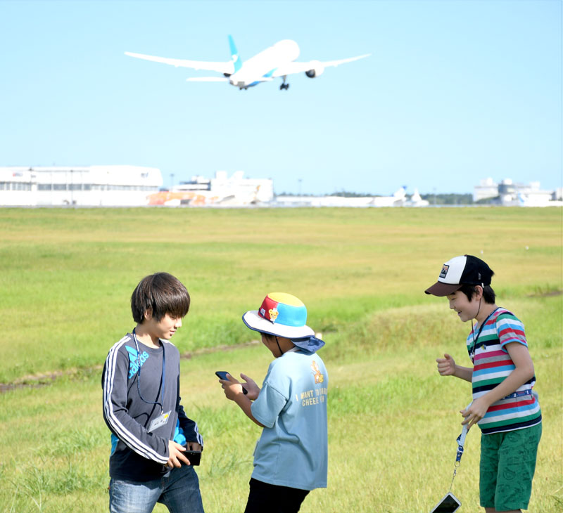 成田空港の滑走路脇から巨大な航空機の離陸シーンを眺める子どもたち