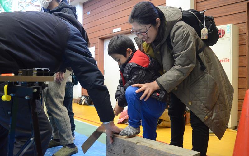 木工品製造体験のブースで、指導を受けながらのこぎりで板を切る男児