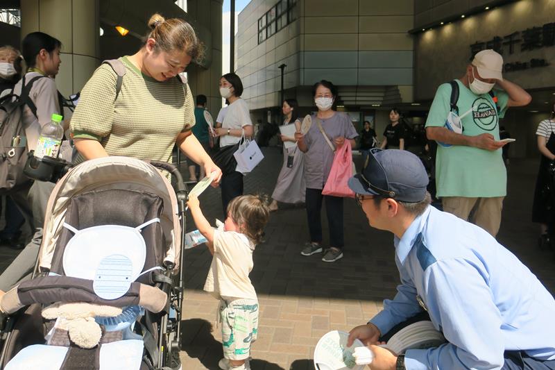 車内放置の危険性を伝えるメッセージ入りうちわを警察官から受け取る親子＝３日、千葉市中央区の京成千葉駅西口前広場