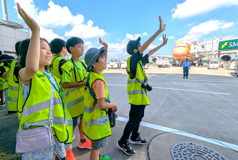 成田空港の駐機場から搭乗橋を歩く旅客に向けて手を振る小学生たち