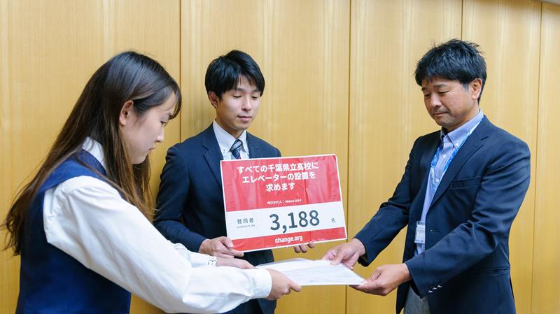 県教委の担当者(右)に集めた署名を提出した松井さん(署名サイトのChange.org Japan提供)=5月、県庁