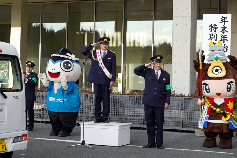 制服にたすき姿でパトロール出発車両を見送る「一日警察署長」の増山さんら＝勝浦市