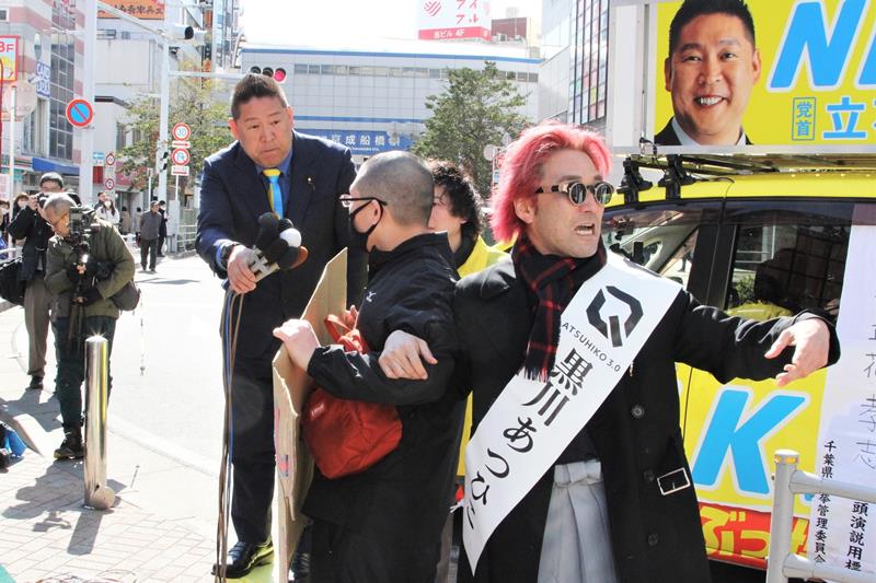 千葉県知事選の第一声で、抗議のプラカードを掲げる聴衆にマイクを向ける立花氏(左)と、聴衆を誘導する黒川氏(右)=2月27日、JR船橋駅前